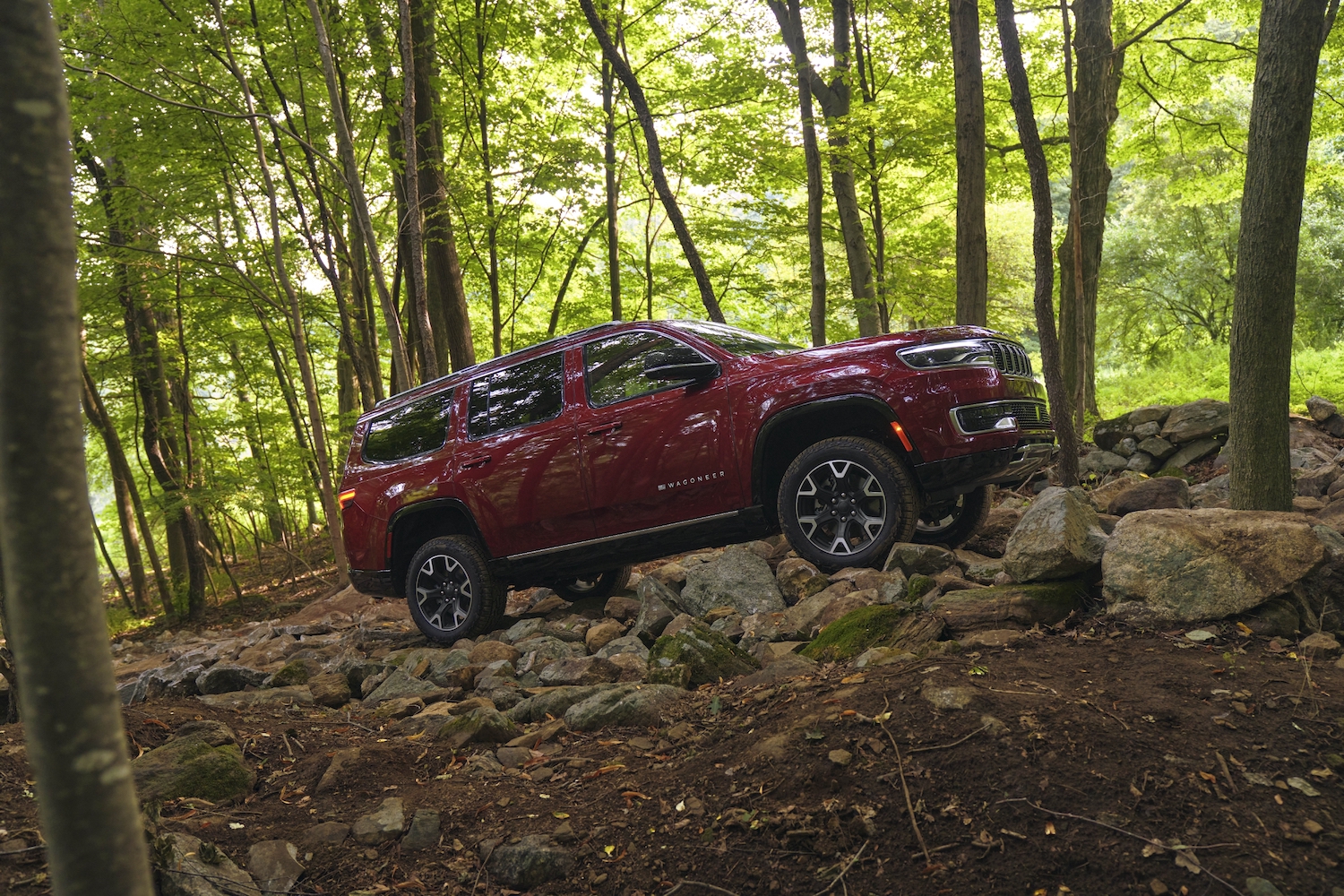 Red Wagoneer SUV climbing a rocky outcropping in the woods, trees visible in the background. 