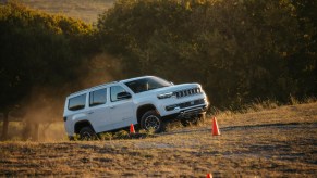 White 2023 Grand Wagoneer SUV climbing a steep hill in a challenge course, a line of trees lit by the sunset visible behind it.