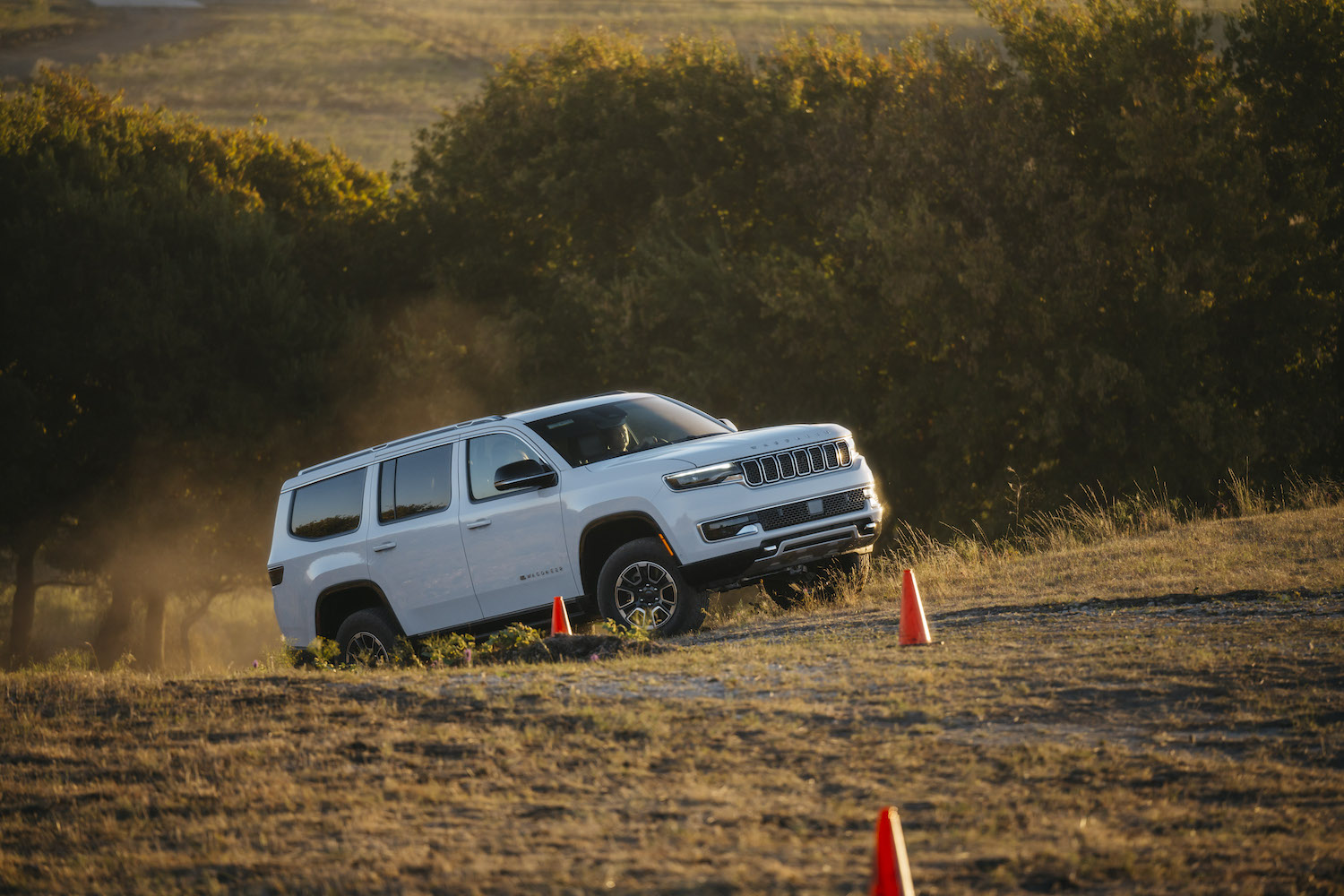 White 2023 Grand Wagoneer SUV climbing a steep hill in a challenge course, a line of trees lit by the sunset visible behind it.