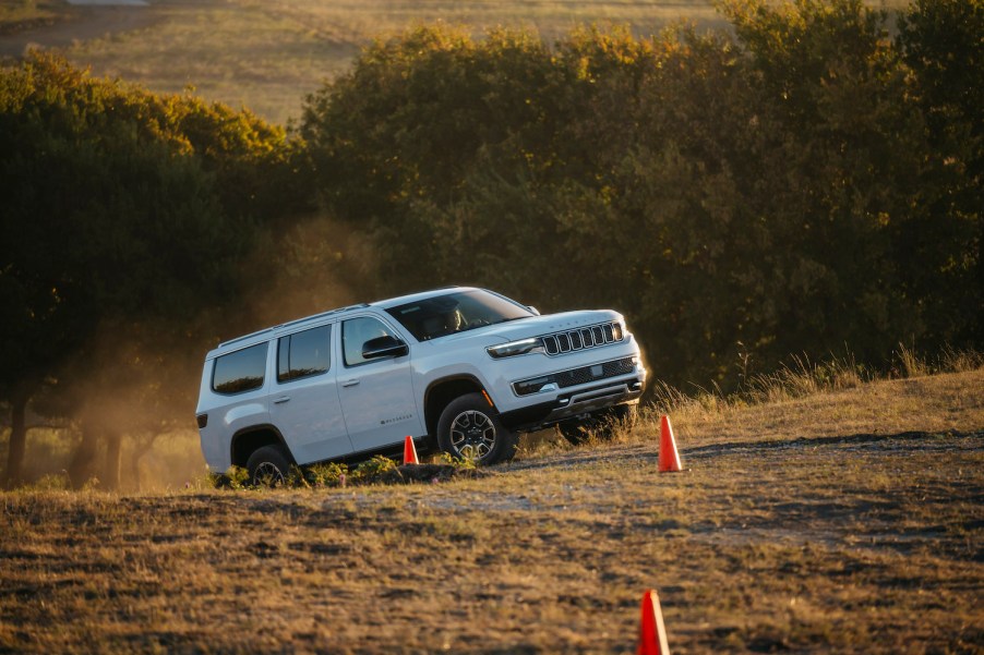 White 2023 Grand Wagoneer SUV climbing a steep hill in a challenge course, a line of trees lit by the sunset visible behind it.