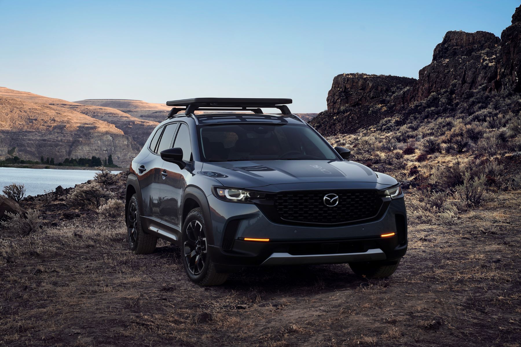 A 2023 Mazda CX-50 compact crossover SUV with roof rails parked in the dirt near water and mountains