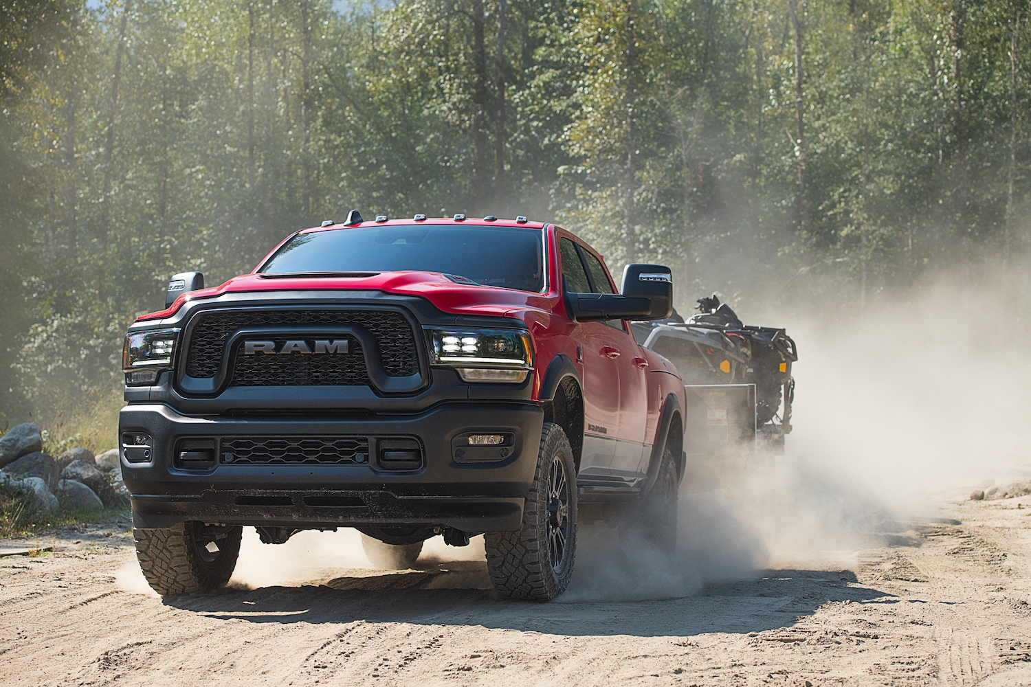 Red Ram 2500 Rebel heavy-duty off-road pickup truck towing a trailer of 4x4s through the woods, a dust cloud visible behind it.