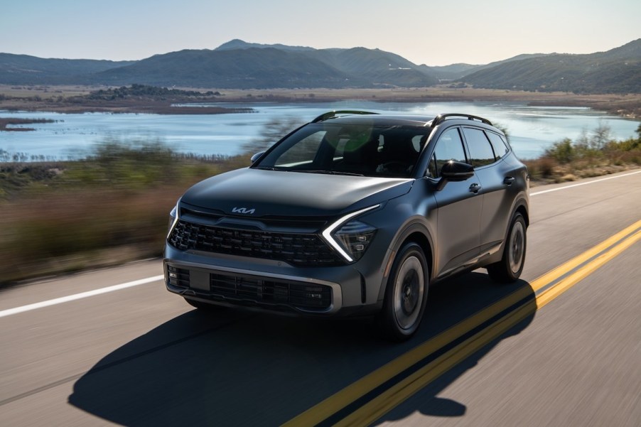 A grey 2023 Kia Sportage PHEV driving down a country road with mountains and water in the background.