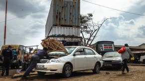 A person at a junk yard, that may want a fixer-upper, looking under the hood of a car.