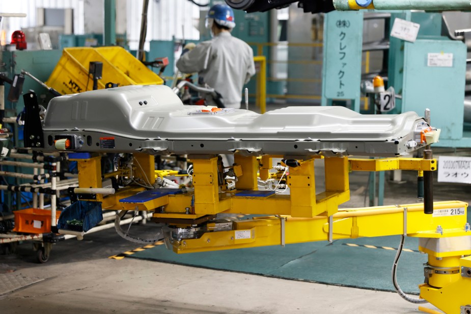 An electric vehicle battery pack in a Mitsubishi Motors factory, destined for a KEI EV, a worker in a hard hat visible in the background.