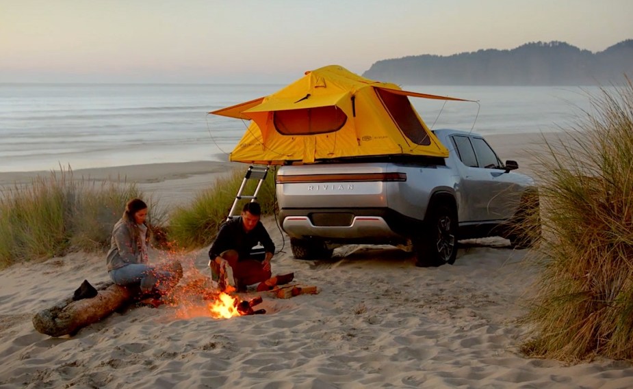 Two people sit by a fire in front of a silver Rivian R1T electric pickup truck parked on a beach.