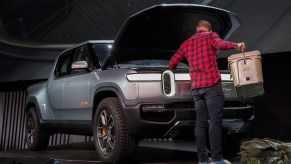 A man setting a cooler in the front trunk of a silver Rivian electric truck parked on a stage.