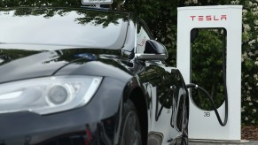 Closeup of the front headlight of a black Tesla electric vehicle charging its Lithium-ion battery at a supercharger visible in the background.