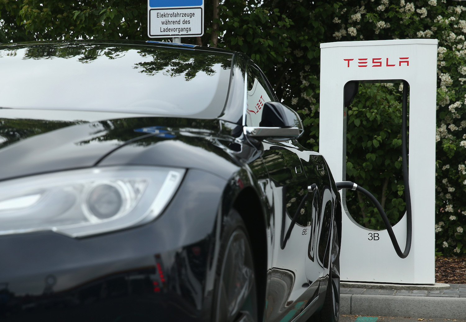 Closeup of the front headlight of a black Tesla electric vehicle charging its Lithium-ion battery at a supercharger visible in the background.