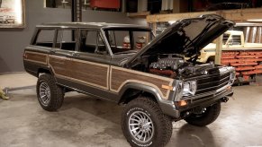 A Jeep Grand Cherokee restomod with a supercharged Hellcat V8 parked in a garage, a pickup truck and tools visible in the background.