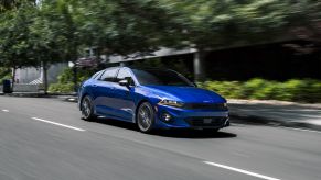 A dark blue 2022 Kia K5 midsize sedan model driving past of line of trees shading a sidewalk