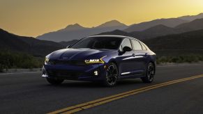 A dark blue 2023 Kia K5 midsize sedan model parked on a country highway at sunrise