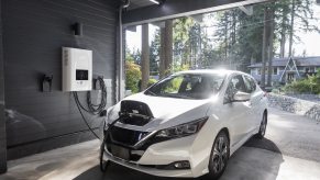 White electric car plugged into a charging cable while parked in a garage, a sunny street visible in the background.