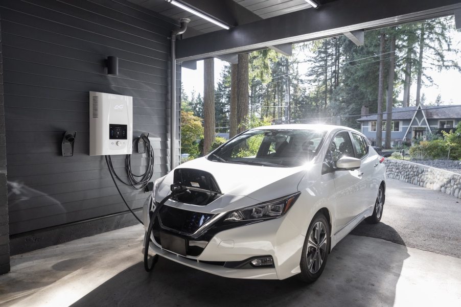 White electric car plugged into a charging cable while parked in a garage, a sunny street visible in the background.