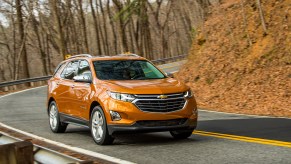 Orange Chevrolet Equinox crossover SUV driving around a curve in a rural road, trees and orange leaves visible in the background.