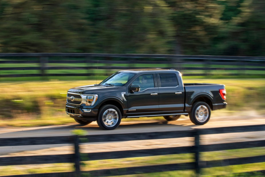 Promo photo of a dark gray Ford F-150 powerboost hybrid truck driving down a country road, a fence visible in the background.