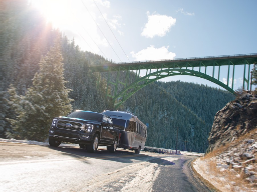 Black Ford F-150 pulls an airstream camper trailer up a steep hill, beneath a bridge, trees visible in the background.