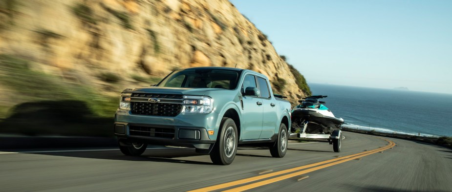 A Ford Maverick hybrid compact pickup truck pulling a jet ski up a steep road, the ocean visible in the background.