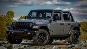 A gray Jeep Wrangler "Willys" edition hybrid 4xe parked on a pile of rocks for a promo photo, trees visible in the background.