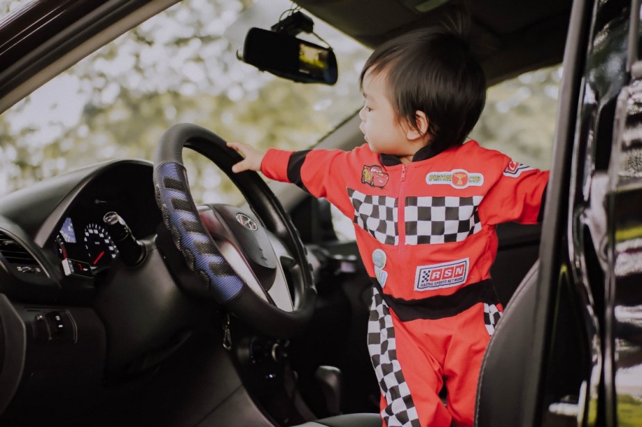 Baby standing by steering wheel of car, highlighting why some babies hate cars seats and how to address it