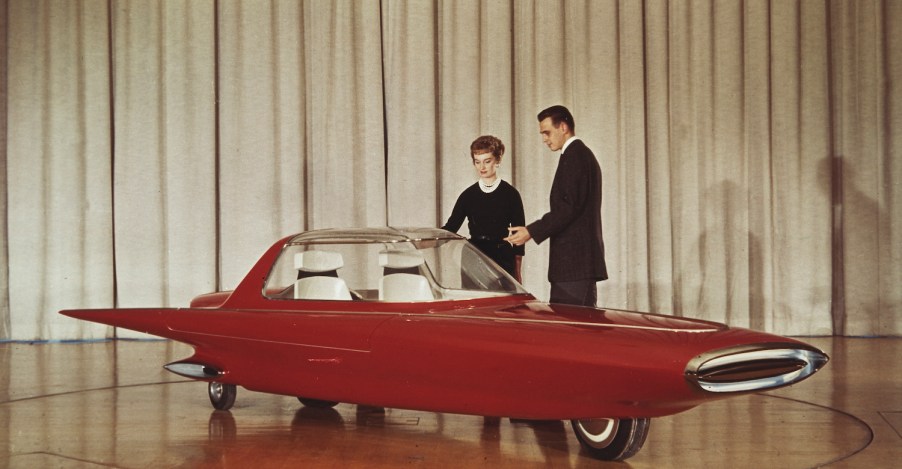 A couple stands next to the 1961 Ford Gyron concept car on stage at the Detroit auto show, a white curtain visible behind them.