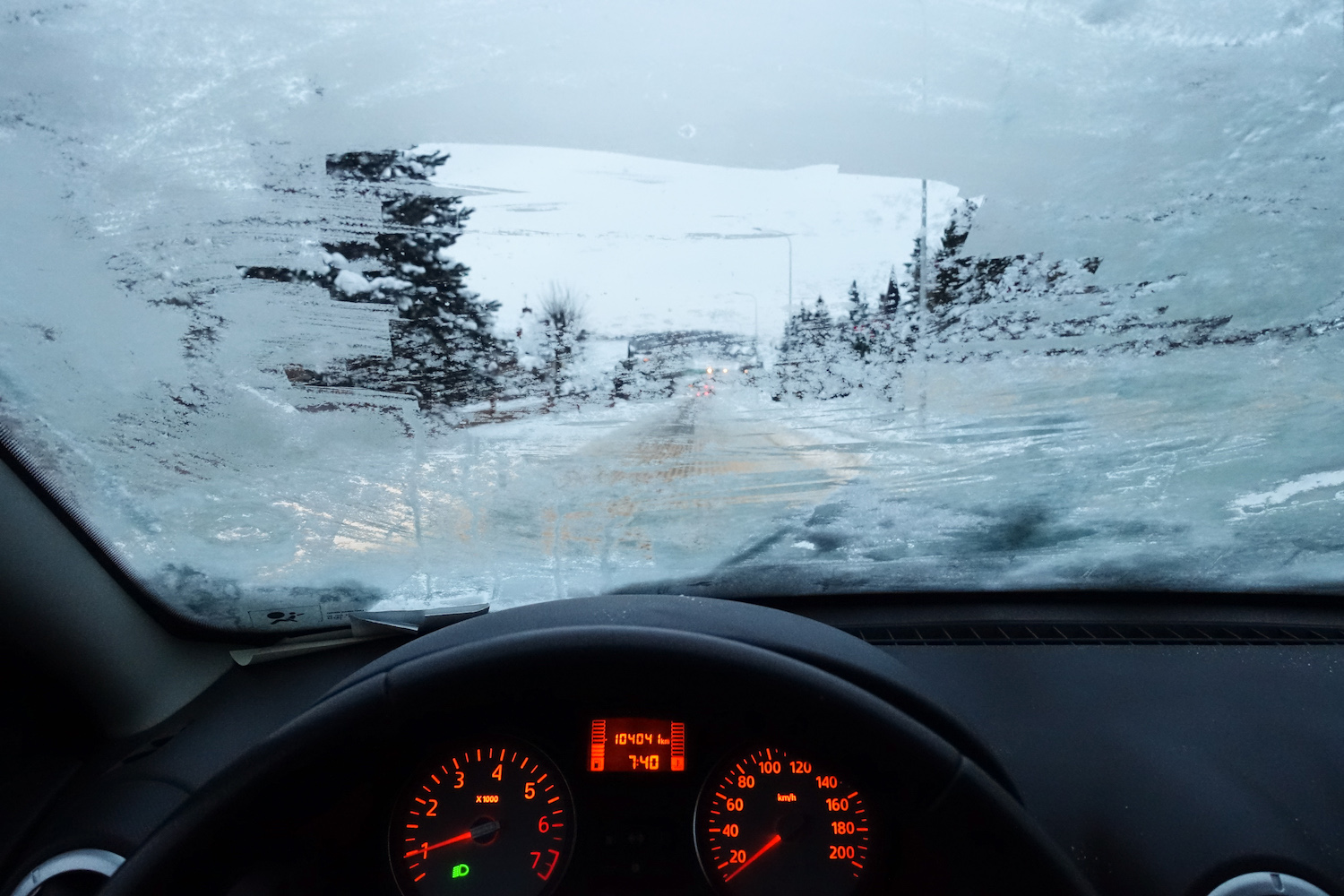 The view of a snowy winter road through a partially defrosted car windshield.