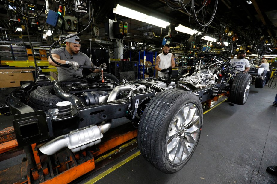 A Ford full-size vehicle chassis built on a ladder frame at a factory assembly line in Kentucky, workers visible bolting on components.