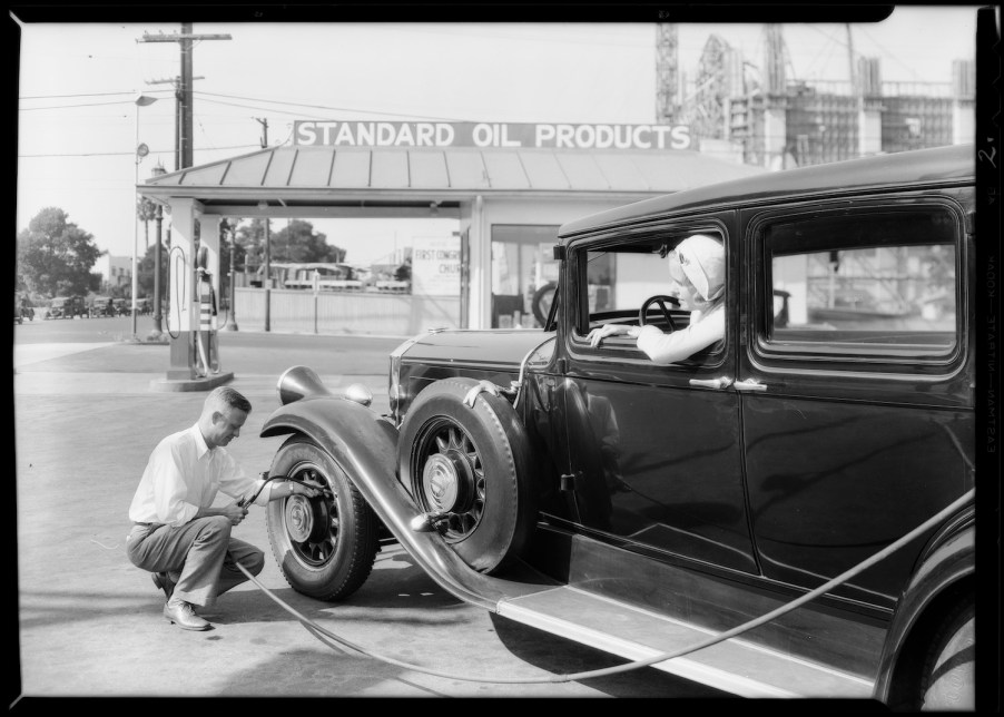 A man crouches by the front tire of a classic car to air up its tires with a hose running from a gas station.