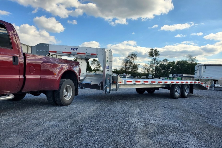 Red dualie pickup truck pulling a flatbed gooseneck EBY trailer through a gravel lot, blue sky visible in the background.