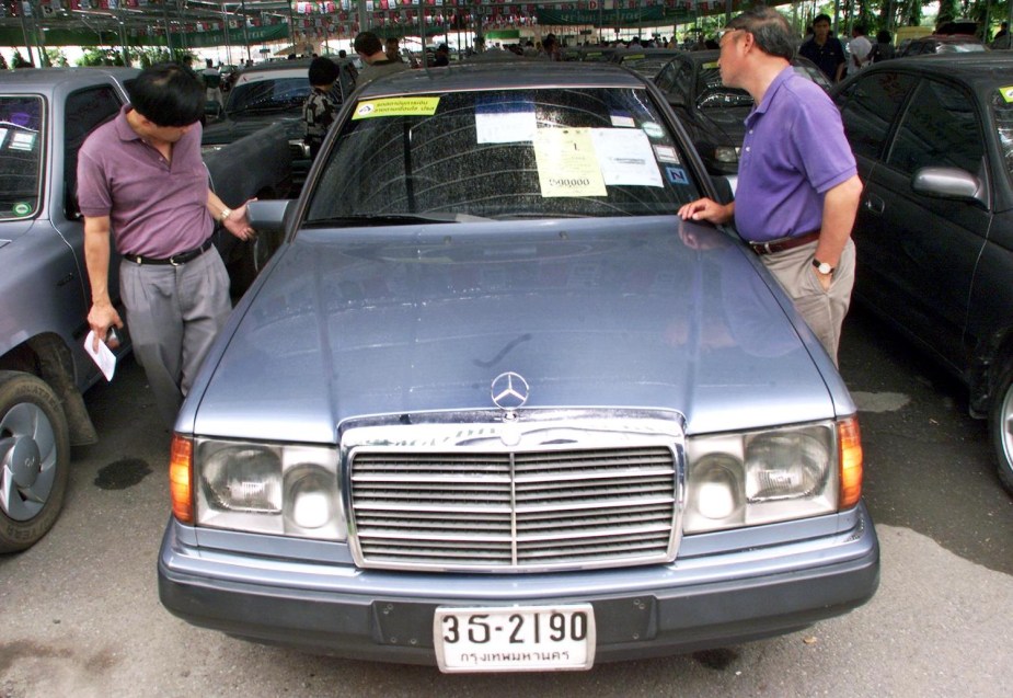 Two men look over a used Mercedes-Benz car during a government auction, its windshield covered in paperwork and other cars visible in the background.