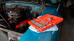 An orange box of sockets and a socket wrench, set on the blue fender of a classic car, its V8 engine visible in the background.