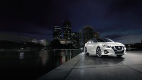 A white 2023 Nissan Maxima midsize sedan parked on a marble plaza near water with a city in the background at night