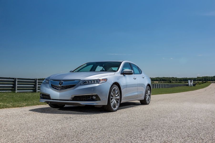 A 2015 Acura TLX entry-level luxury sedan model parked on a gravel road near metal fencing on a farm. The TLX is one of the most reliable Acura models