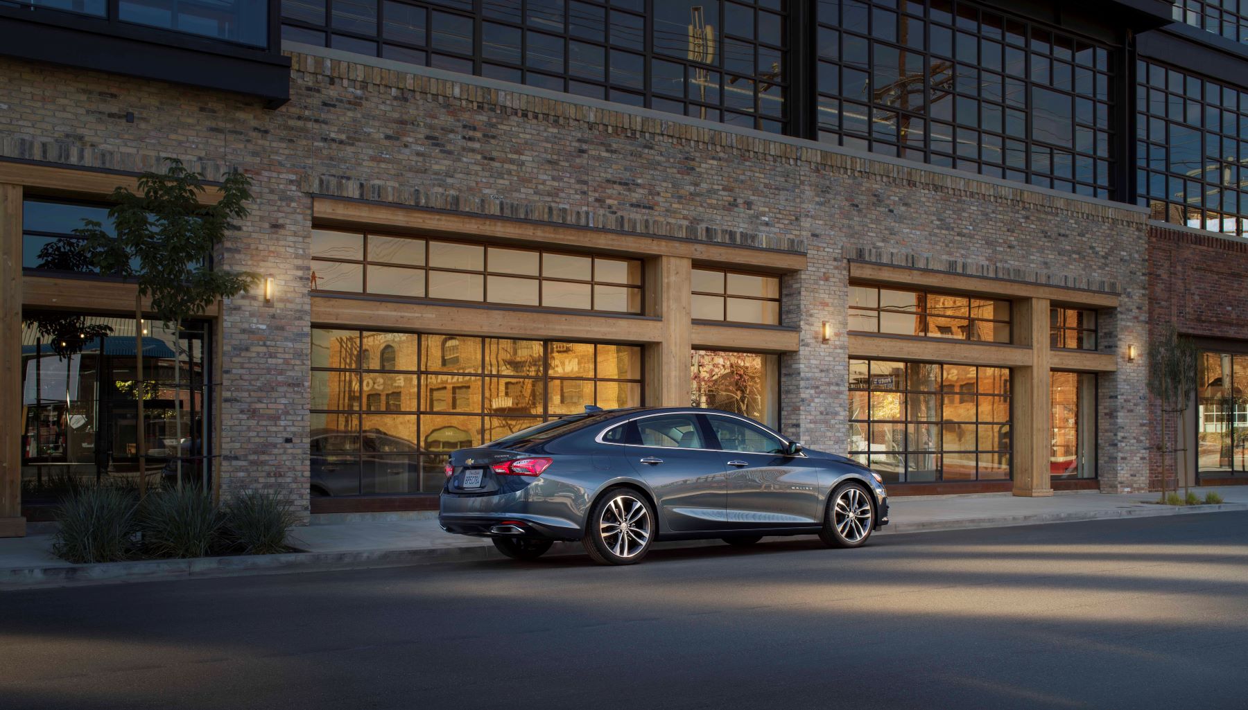 A dark-gray 2021 Chevrolet Malibu midsize sedan model parked outside the windows of a brick building at sunset