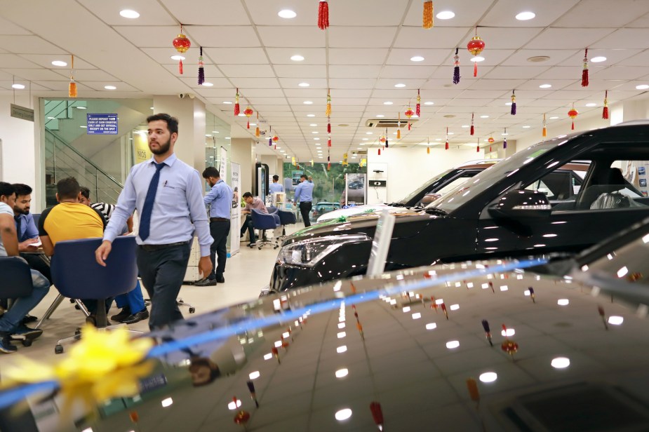 The hood of an EV parked inside a dealership, a table full of customers discuss a potential federal tax credit rebate in the background.