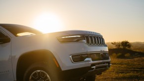 The sunset behind the fender and hood of a white Grand Wagoneer L SUV driving off-road.