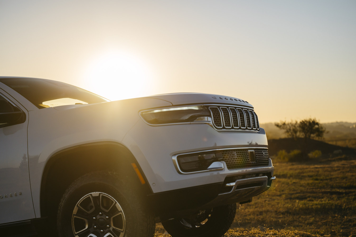 The sunset behind the fender and hood of a white Grand Wagoneer L SUV driving off-road.