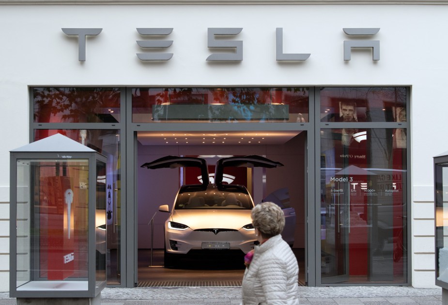 A Tesla model 3 parked in a showroom, a potential buyer visible in the foreground.