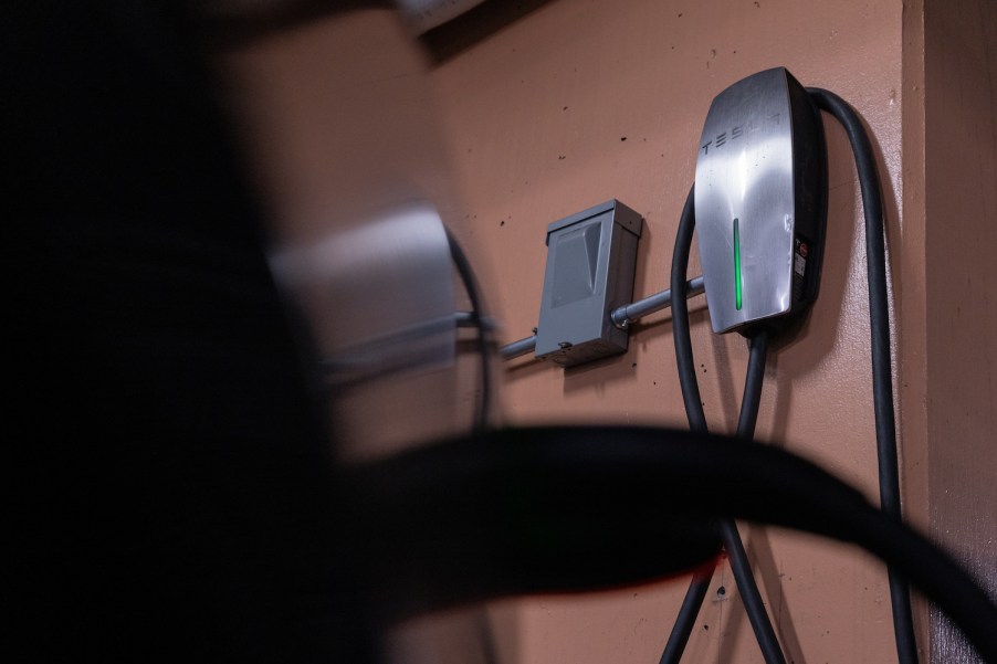 A charging cable plugged into an EV port, a Tesla home charger attached to the wall visible in the background.