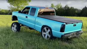 A turquoise-colored squatted pickup truck parked in a field, trees visible in the background.