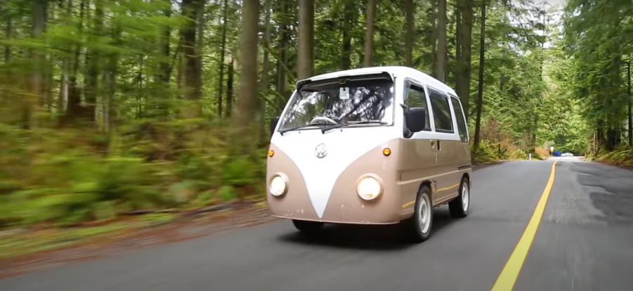 A brown Subaru Sambar converted into a Volkswagen bus lookalike drives on a rural road through pine tree forests.