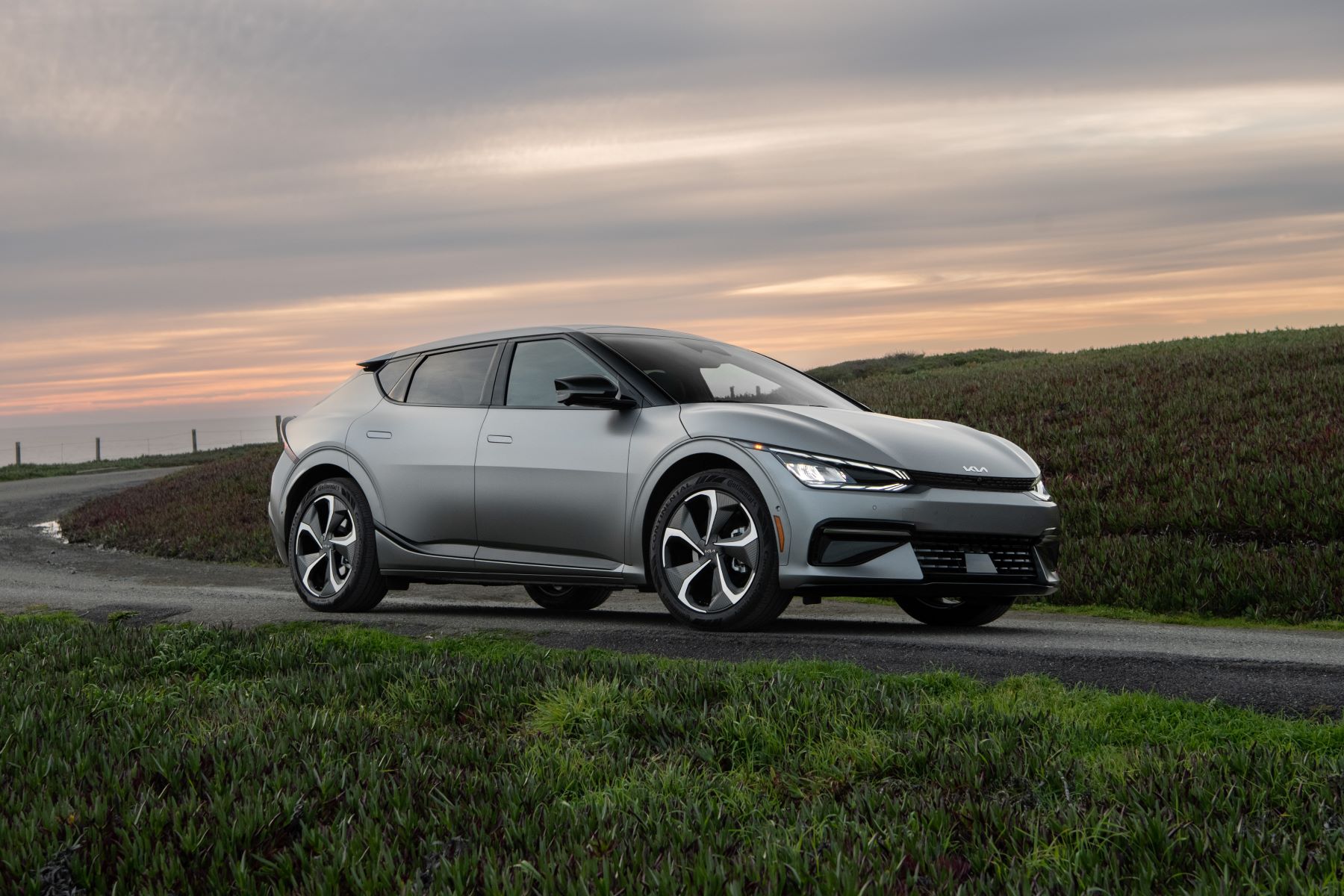 A silver-gray 2023 Kia EV6 compact electric SUV model parked on an asphalt road near a grass field at sunset