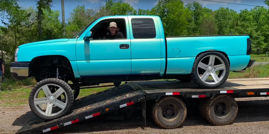 A squatted truck owner drives his pickup off of a travel trailer, trees visible in the background.