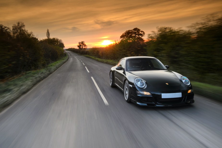 A modified Porsche 997 Carrera S sports car driving on a rural road with a sunset visible in the background.