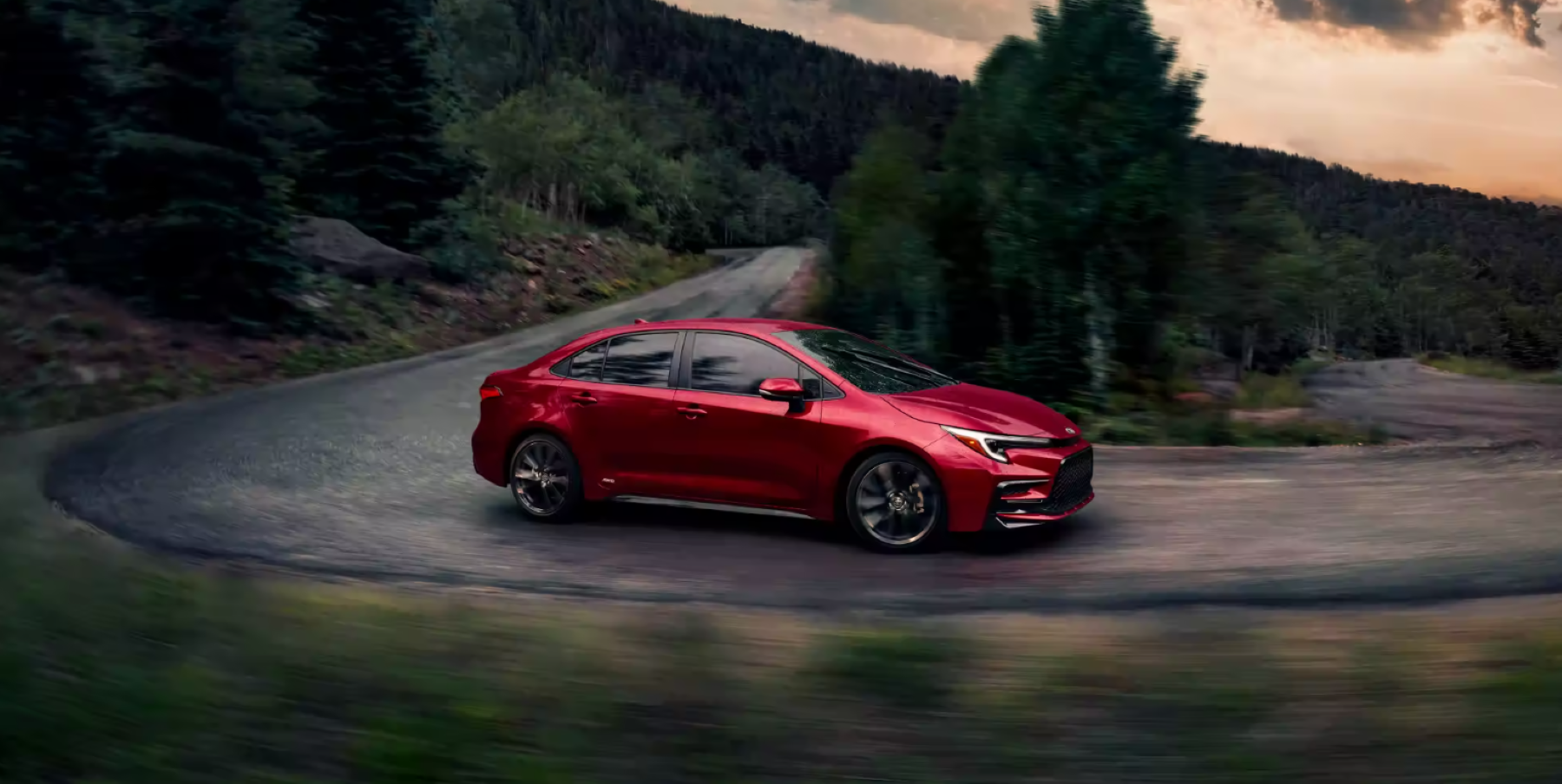 A red 2023 Toyota Corolla Hybrid compact sedan model driving on a curving forest road