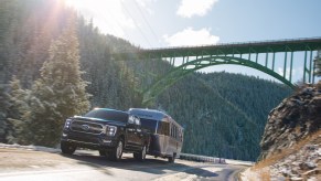 A black Ford F-150 PowerBoost hybrid pickup shows off its truck of the year powertrain by pulling a heavy camper trailer up a steep hill, a bridge visible in the background.