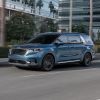 A light blue 2023 Kia Carnival MPV/minivan model driving past glass skyscrapers and greenery