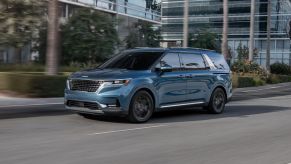 A light blue 2023 Kia Carnival MPV/minivan model driving past glass skyscrapers and greenery