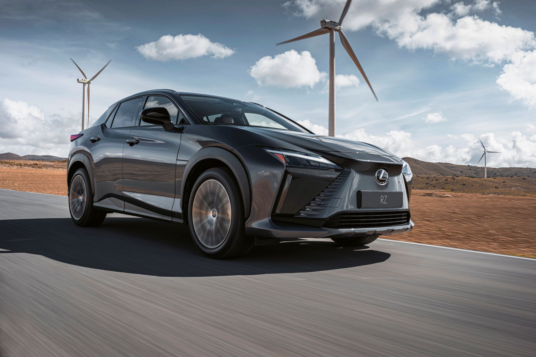 A dark-gray 2023 Lexus RZ all-electric luxury SUV driving on a highway past windmills