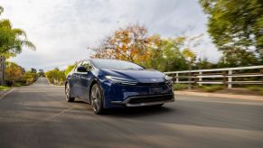 A 2023 Toyota Prius Limited in Reservoir Blue driving past white fencing on a country asphalt road